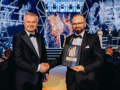 Středně dlouhý záběr dvou mužů ve formálním oblečení s černou kravatou, jak si podávají ruce. Muž vpravo Maciej Góralski drží zarámované ocenění se zlatým emblémem, které je označeno jako cenu „Podnikatel roku 2022“. V rozmazaném pozadí je vidět pódium s abstraktními geometrickými strukturami a dynamickými modrými a červenooranžovými grafickými vzory. Několik hostů sedí u stolů v tlumeně osvětleném sále.