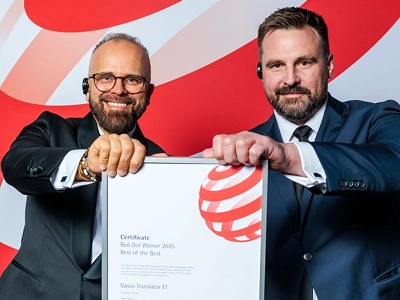 Dva muži ve formálním oblečení – levý ve smokingu s brýlemi, pravý v tmavě modrém obleku – společně drží oběma rukama zarámovaný certifikát. Na certifikátu je uvedeno „Red Dot Winner 2025 Best of the Best“ udělené překladači „Vasco Translator E1“. V pozadí je vidět rozsáhlé, dynamické grafické pozadí s abstraktními červenobílými vlnovými vzory.