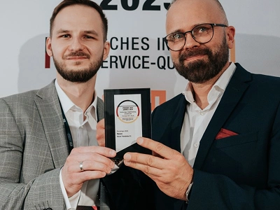 Širokoúhlá fotografie zachycuje pódium pro slavnostní předávání cen zpoza sedícího publika. Na levé straně pódia stojí muž v tmavém obleku a žena v dlouhém bílém županu s tablety v rukou, otočení k davu. Uprostřed tmavého pozadí pódia se na velké obrazovce nahoře zobrazuje zářící jantarový text s nápisem „WIZJA I INNOWACJE“. Pod ním, uprostřed obrazovky, se nachází velké, zářící křišťálové ocenění ve tvaru diamantu, za nímž následuje text „Maciej Góralski“ a pod ním „Vasco Electronics“. Teplé, rozptýlené světlo osvětluje pódium proti tmavému, přeplněnému hledišti.