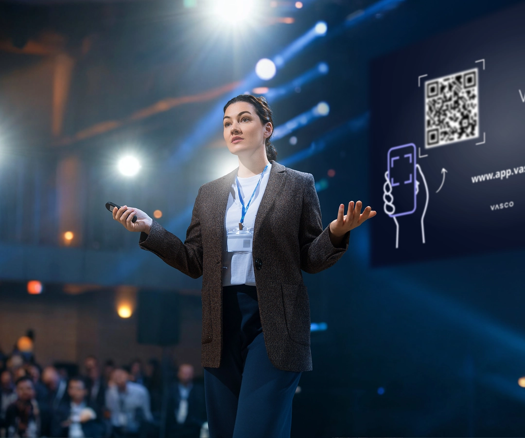 A woman stands on a brightly lit conference stage, presenting to an audience. She wears a brown blazer, a white t-shirt, and a conference ID lanyard. She holds a presentation clicker in her right hand and gestures with her left. Behind her, a large screen displays a prominent QR code for the 'Vasco Assistance' service and a small illustration of a smartphone scanning it. The audience is blurred in the foreground.