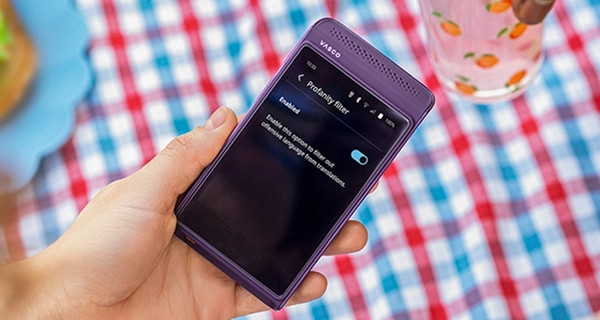 A close-up photograph of a hand holding a purple Vasco electronic device. The device's screen displays the "Profanity filter" settings menu, showing the feature is "Enabled" with a blue toggle switch turned on and text that reads, "Enable this option to filter out offensive language from translations." The background features a red, white, and blue checkered tablecloth, with another hand reaching for a clear glass decorated with peach illustrations in the upper right corner.