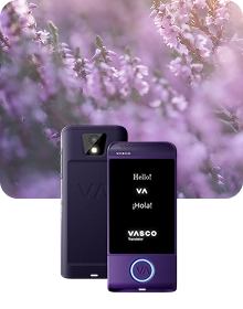 Prezentační fotografie produktu zachycující fialové zařízení Vasco Translator ze dvou úhlů na pozadí tmavé texturované látky. Zařízení vlevo je zobrazeno zezadu, přičemž je zdůrazněn objektiv fotoaparátu s bleskem a reliéfní logo „VA“. Zařízení vpravo je zobrazeno zepředu s rozsvíceným displejem, na kterém se zobrazují pozdravy „Hello!“, „VA“, „¡Hola!“ a logo „VASCO Translator“. Kruhové tlačítko ve spodní části přední strany zařízení je orámováno zářícím modrým světlem. Kompozice je zasazena do snového, rozmazaného pozadí fialových květů vřesu zalitých měkkým slunečním světlem.