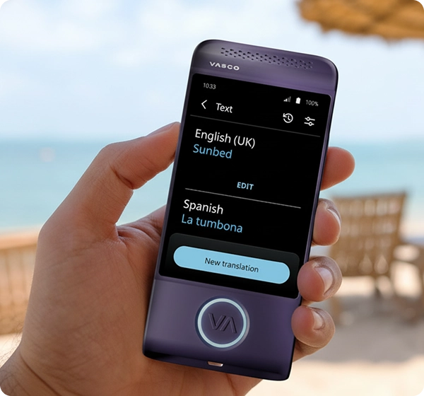 Detailní záběr ruky, která drží fialový elektronický překladatel značky Vasco. Na displeji je zobrazen překlad textu z angličtiny (britská varianta) do španělštiny, kde je slovo „Sunbed“ přeloženo jako „La tumbona“. Pozadí tvoří jasná, slunná plážová scéna s rozostřeným výhledem na oceán, zlatý písek, několik lehátek a slaměný slunečník. 